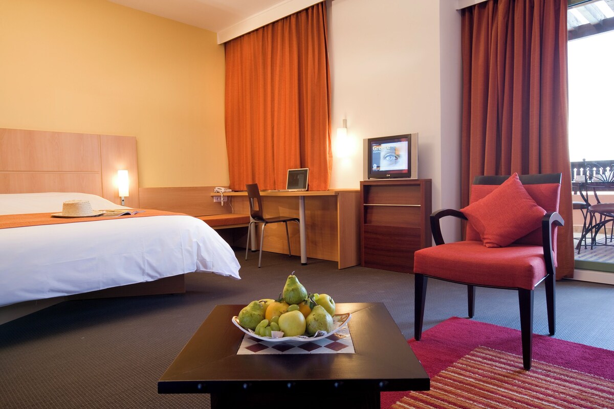 A hotel room with a bed, desk, chair, and a table with fruit.
