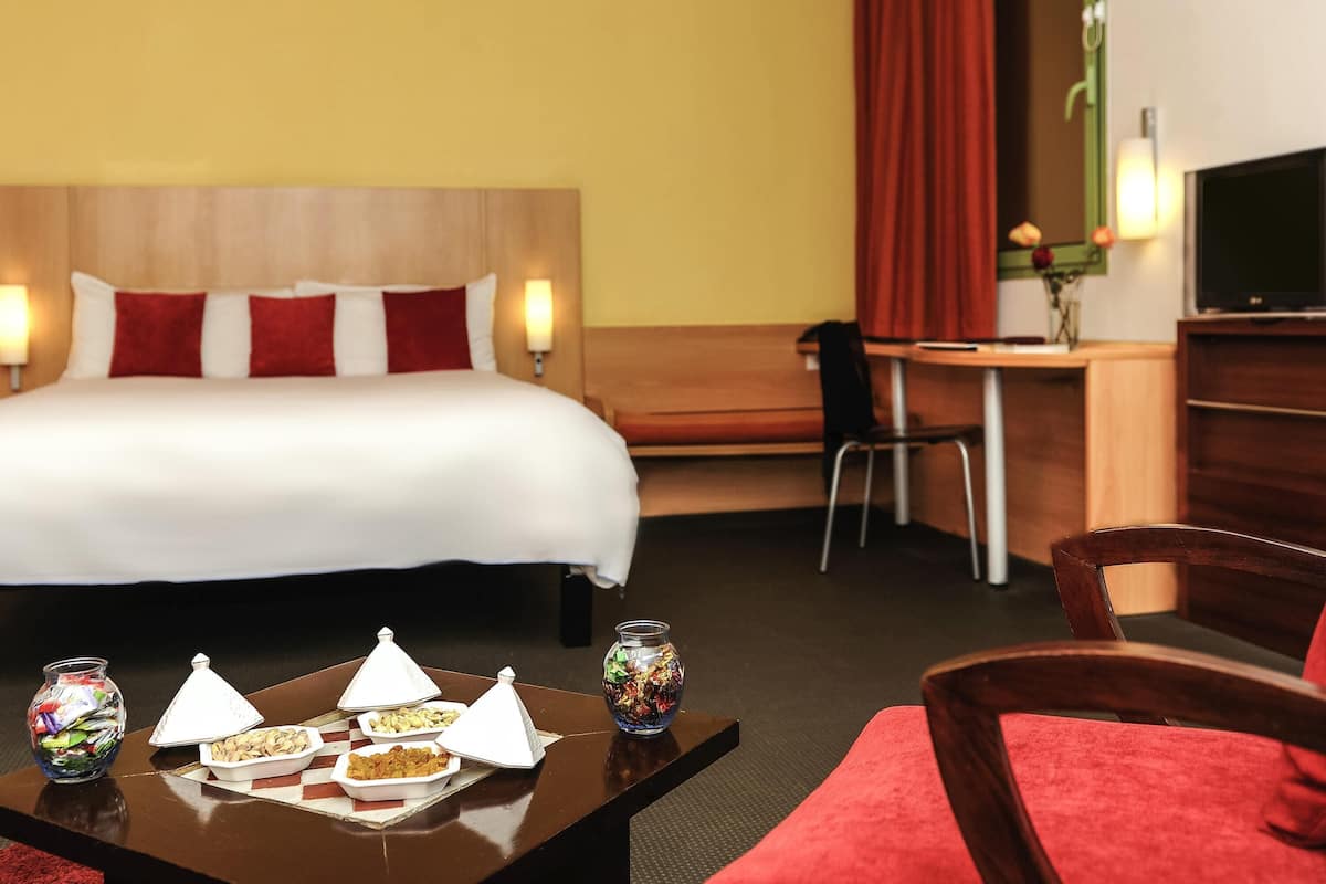 A hotel room with a bed, desk, chair, TV, and a table with food and a decorative bowl.