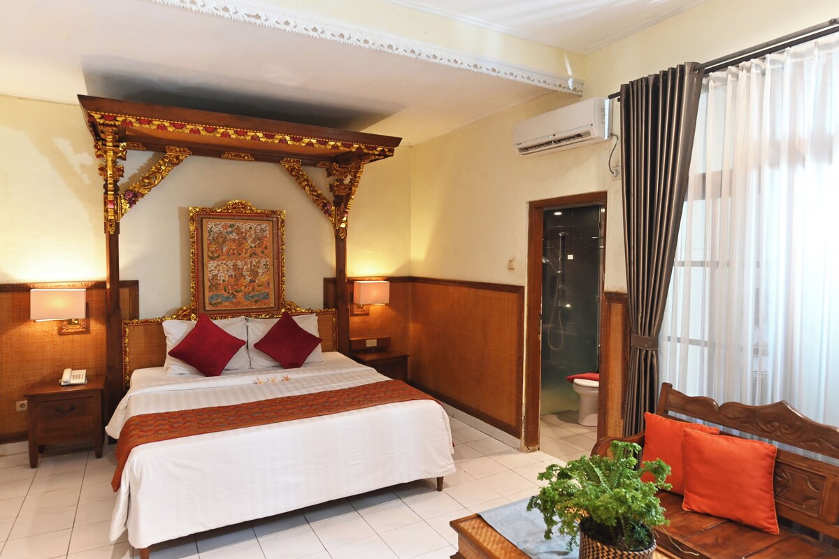 Habitación de hotel con una cama grande, un dosel de madera, una obra de arte enmarcada sobre la cama, un banco de madera con cojines y un equipo de aire acondicionado en la pared.