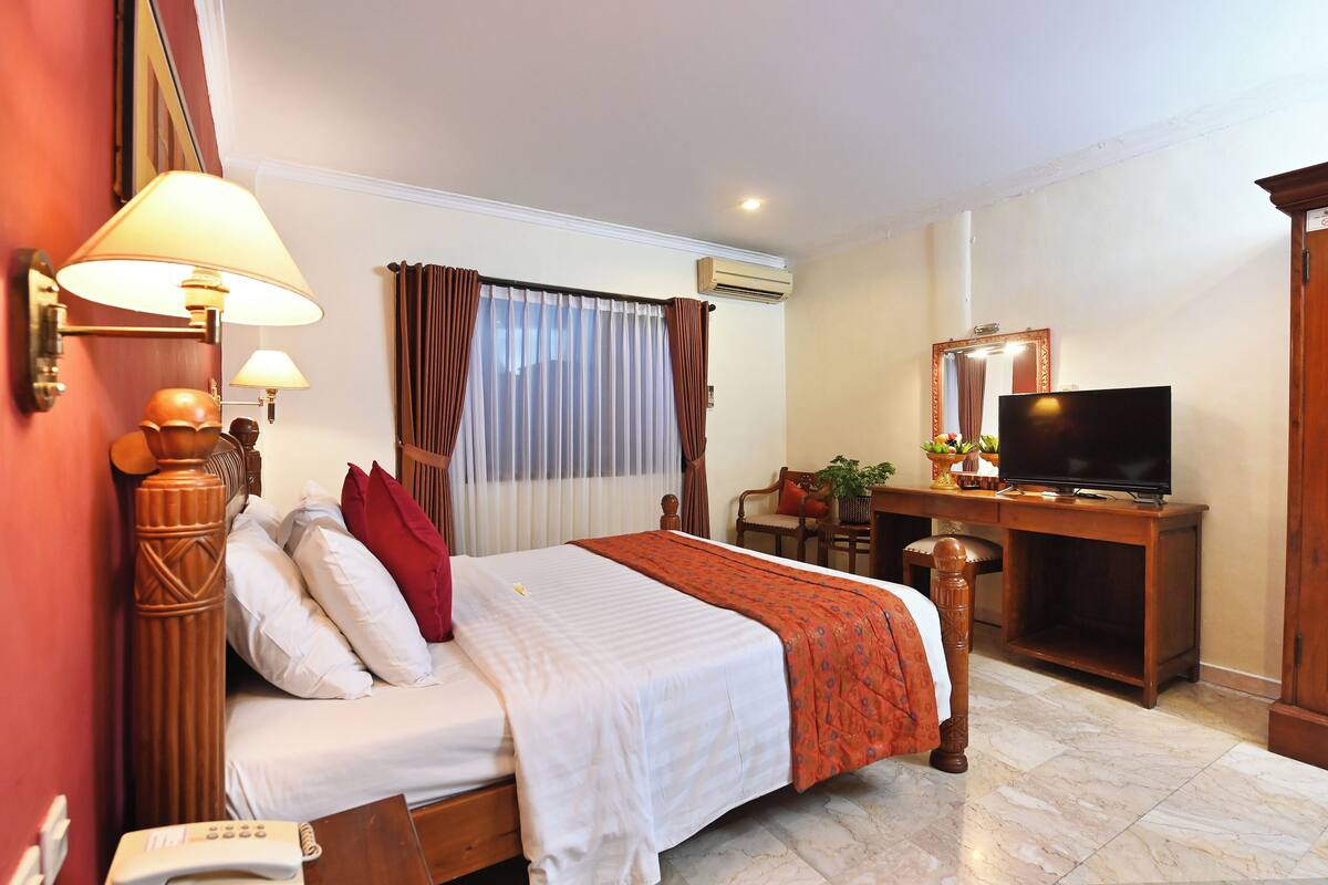 A hotel room with a bed, bedside lamps, a desk with a television, and an air conditioning unit.