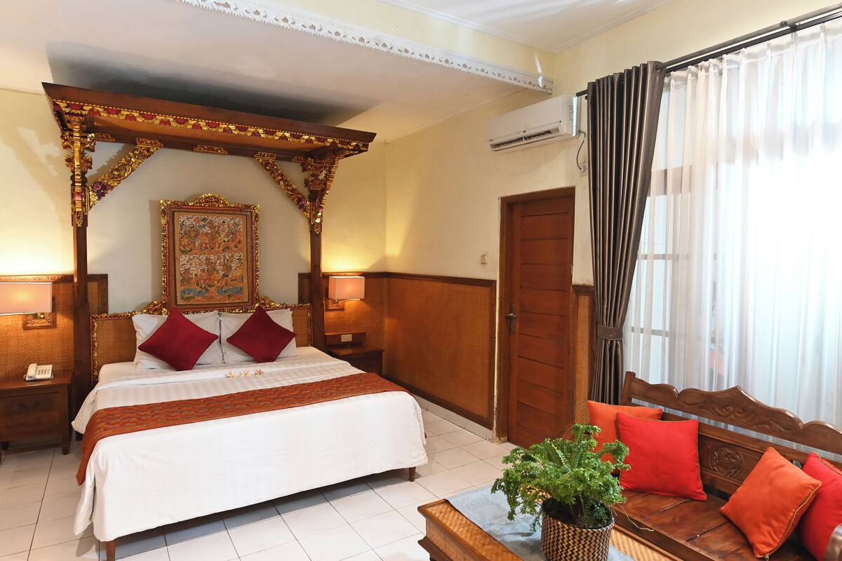 A hotel room with a large bed, a wooden canopy, a wooden bench with red cushions, and a potted plant.