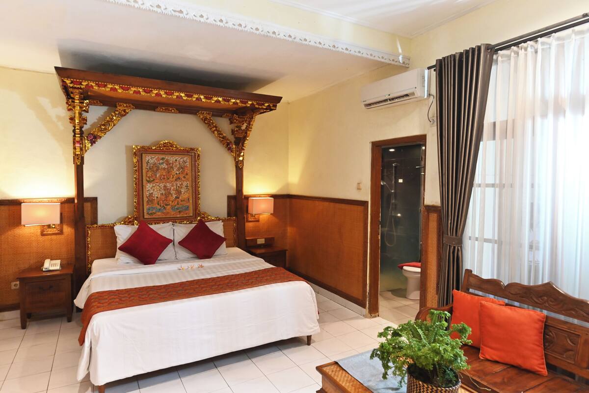 A hotel room with a large bed, a wooden canopy, a framed artwork above the bed, a wooden bench with cushions, and an air conditioning unit on the wall.