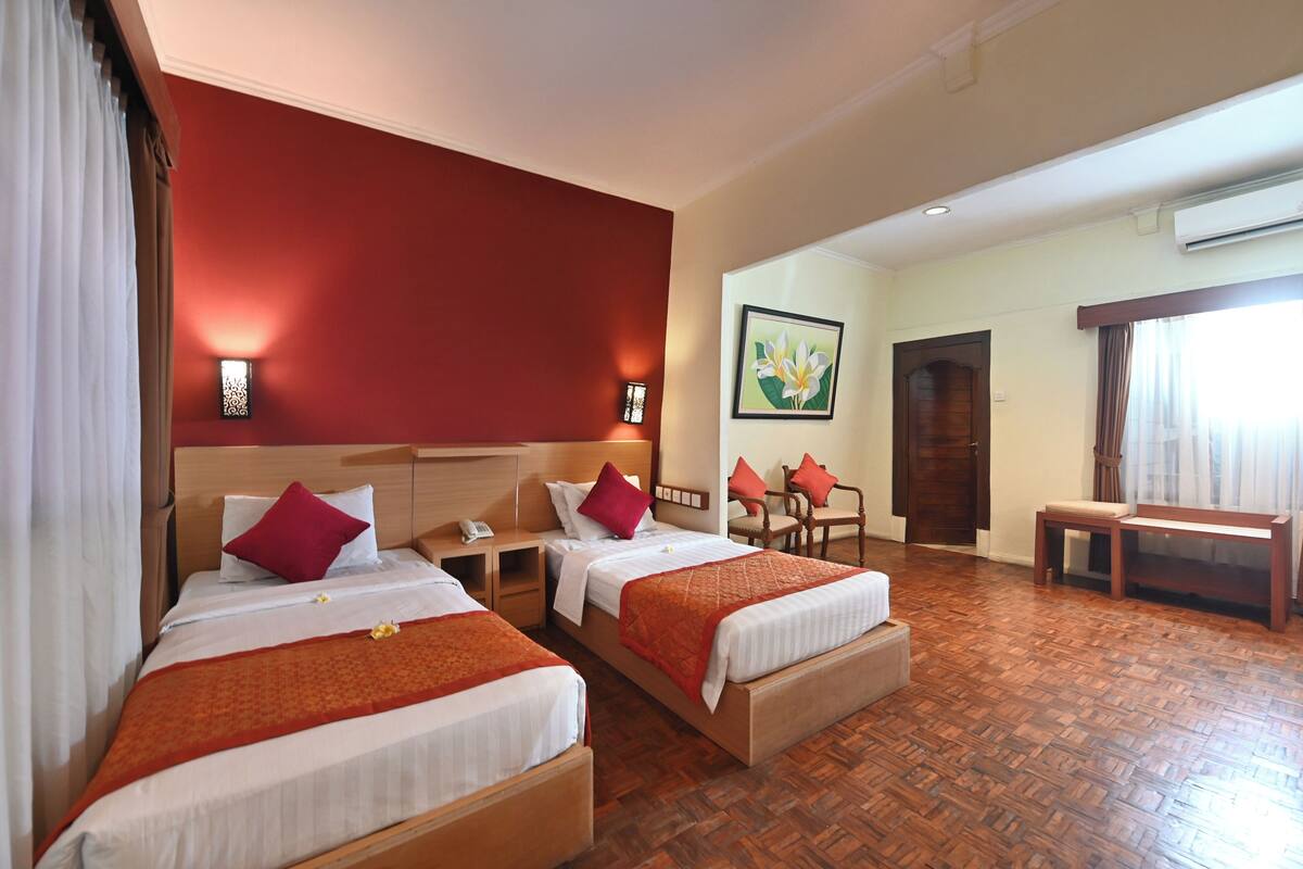 A hotel room with two beds, a wooden floor, a desk, a chair, a bench, and a painting on the wall.