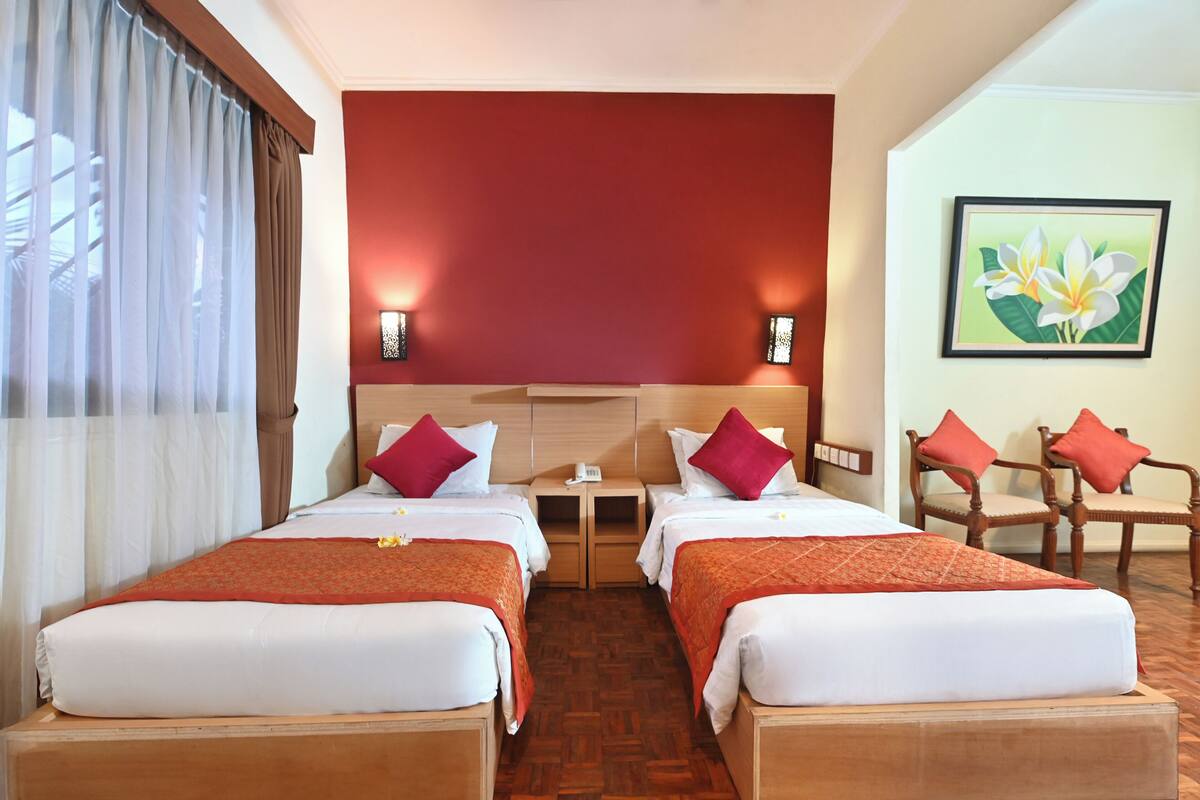 A hotel room with two beds, a wooden headboard, a small table, two wall-mounted lamps, and a painting of flowers.