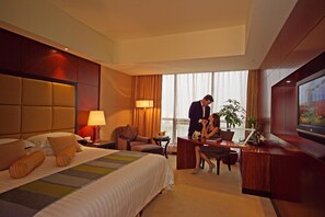Ein Hotelzimmer mit einem großen Bett, einem Schreibtisch, einem Sessel, einem Fernseher und einem Fenster mit Vorhängen.