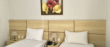 A hotel room with two beds, each with a white bedspread and pillows, a wooden headboard, bedside tables with lamps, and a wall-mounted painting of flowers.