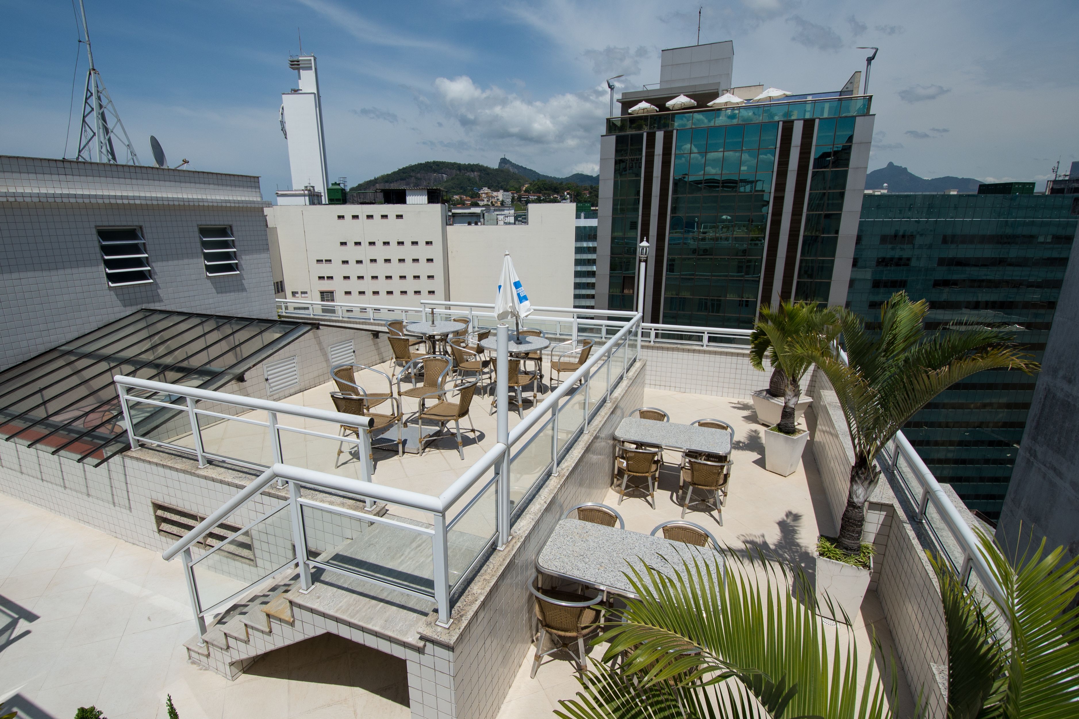 A rooftop terrace with outdoor seating, a glass balustrade, and a view of the cityscape and mountains.