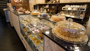A bakery counter with various pastries and desserts displayed in a glass case, a coffee machine, and shelves stocked with bottles and condiments. - Hotel OK (Rio de Janeiro)
