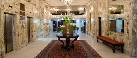 A marble lobby with a chandelier, a round table with flowers, and a bench.