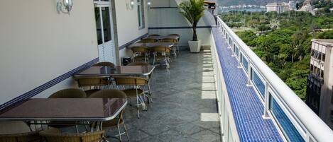 A balcony with tables and chairs, a potted plant, and a view of the city and mountains.