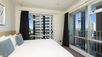 Habitación de hotel con una cama grande, vistas a un paisaje urbano y un balcón con barandilla de vidrio.