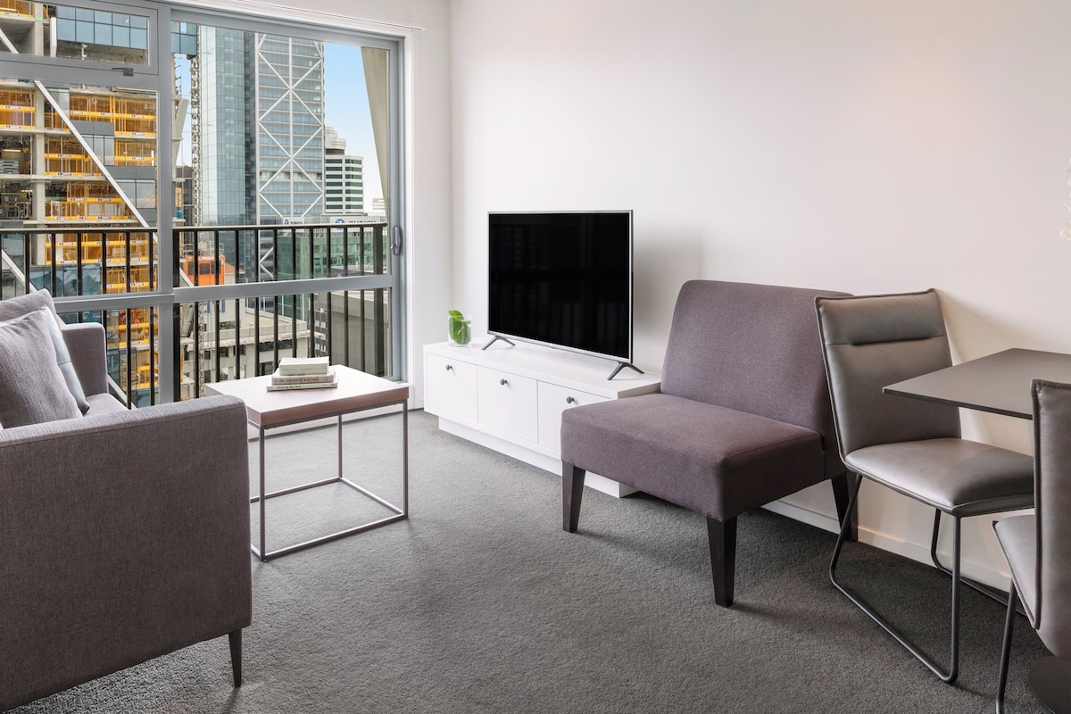 A modern living room with a sofa, armchair, and a small table. A TV is mounted on the wall, and there is a large window with a view of a cityscape.