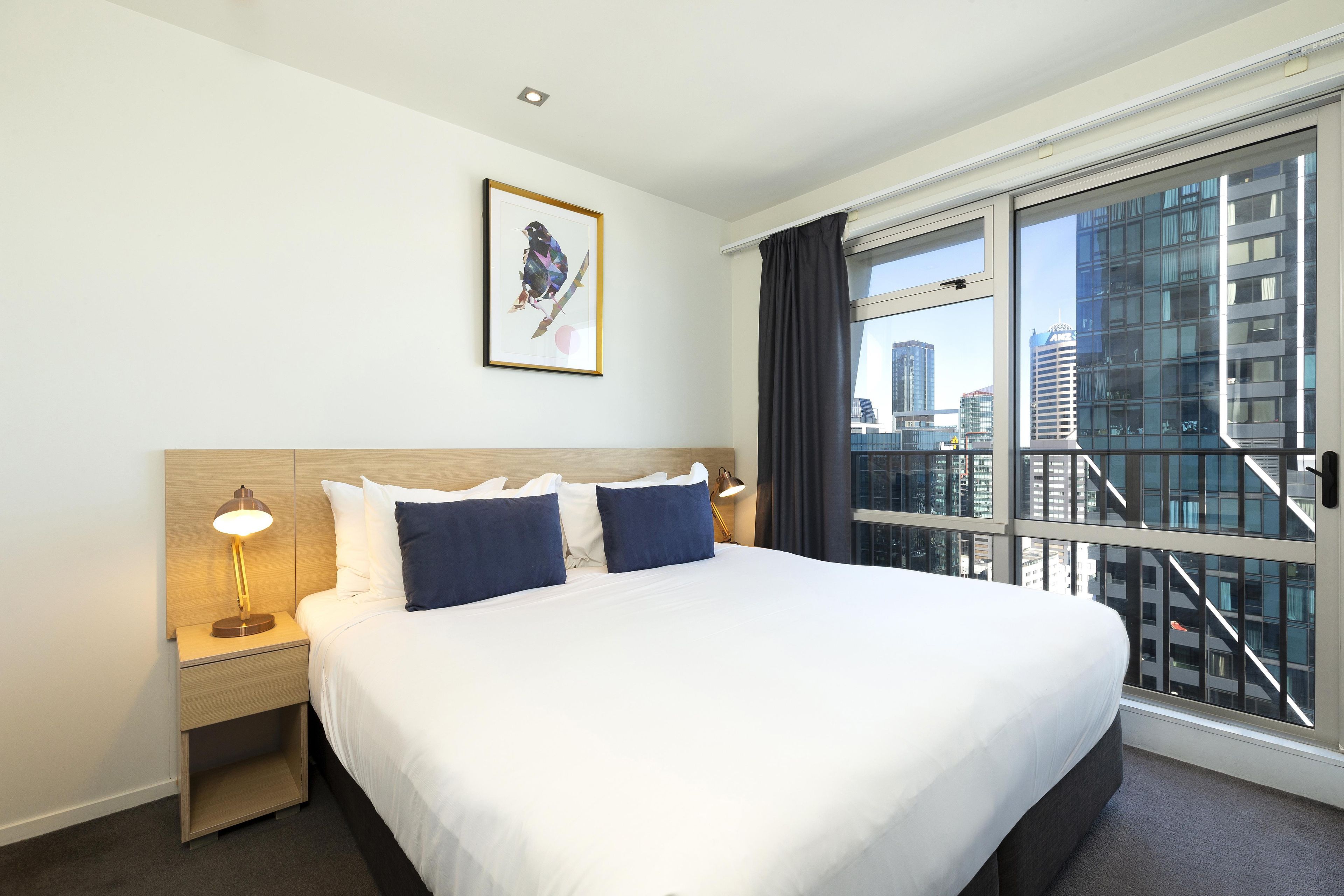 A hotel room with a large bed, two bedside tables with lamps, a framed artwork on the wall, and a view of a cityscape through a large window.