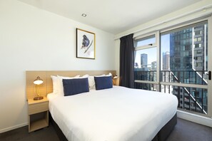 A hotel room with a large bed, two bedside tables with lamps, a framed artwork on the wall, and a view of a cityscape through a large window. - Auckland Harbour Suites (Auckland)