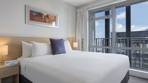 A hotel room with a large bed, a nightstand with a lamp, a framed picture on the wall, and a view of the city through large windows. - Auckland Harbour Suites (Auckland)
