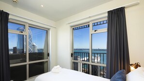 A hotel room with a bed, a window with a cityscape view, and a balcony with a railing. - Auckland Harbour Suites (Auckland)