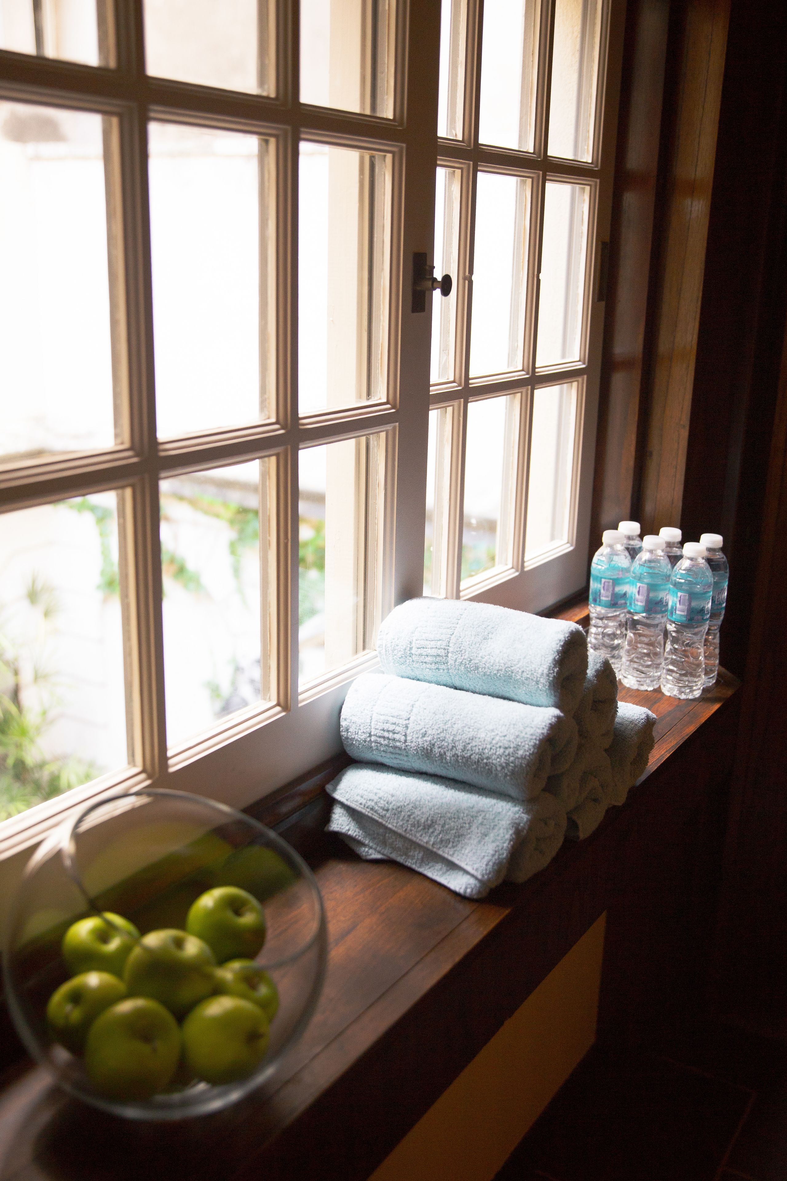 A window with a view of greenery, a bowl of green apples, and several water bottles on a wooden ledge.