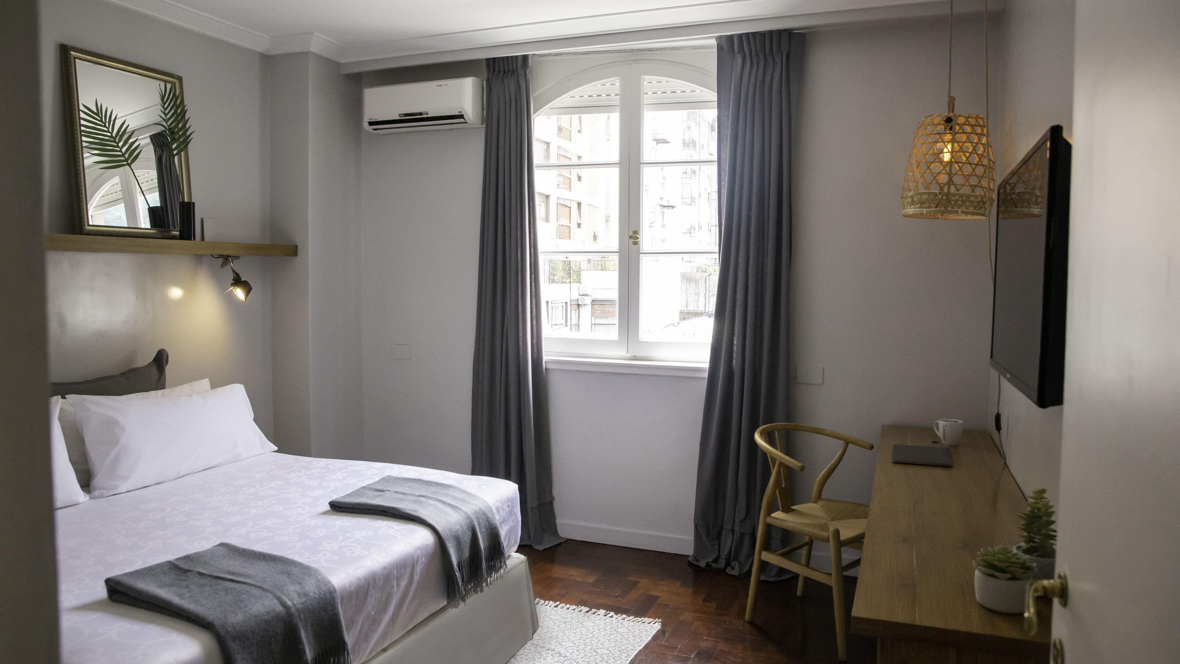 A bedroom with a bed, a desk, a chair, a TV, a window with curtains, and a plant.