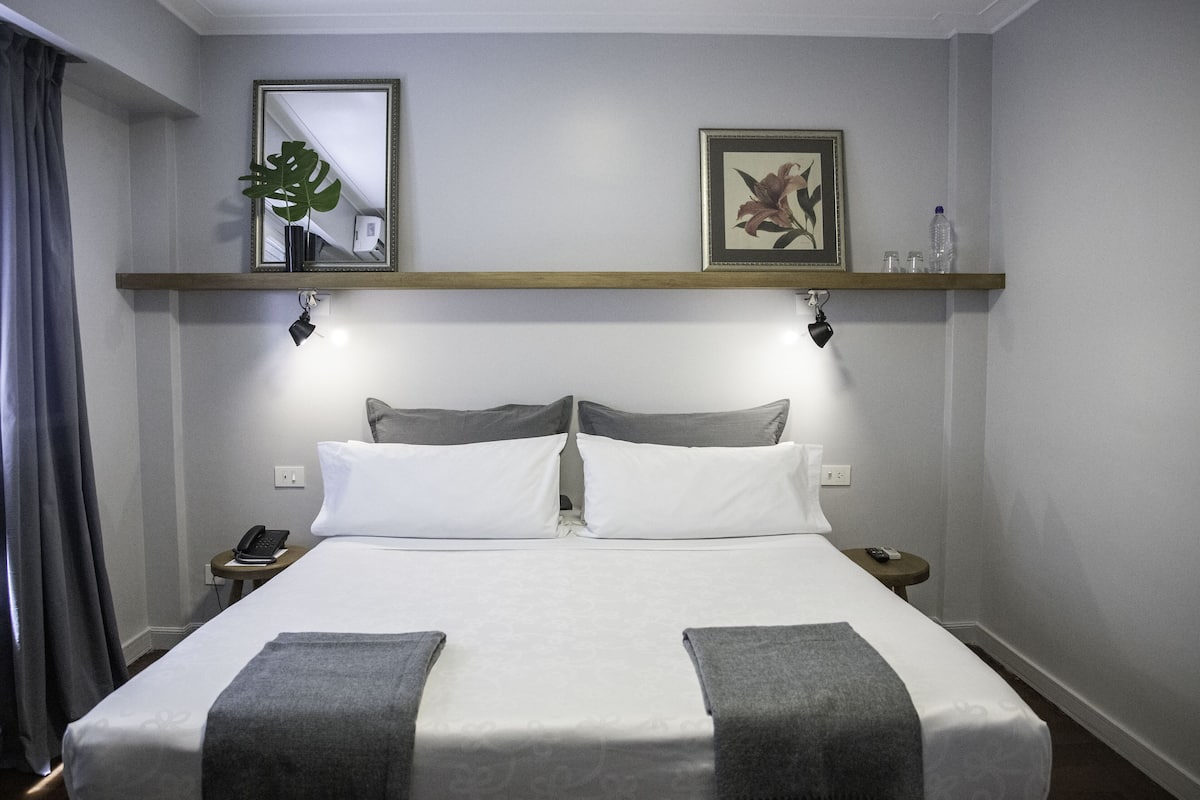 Een keurig opgemaakt bed met een grijs-witte spreideken, twee nachtkastjes met lampen, een spiegel en een ingelijste foto aan de muur.