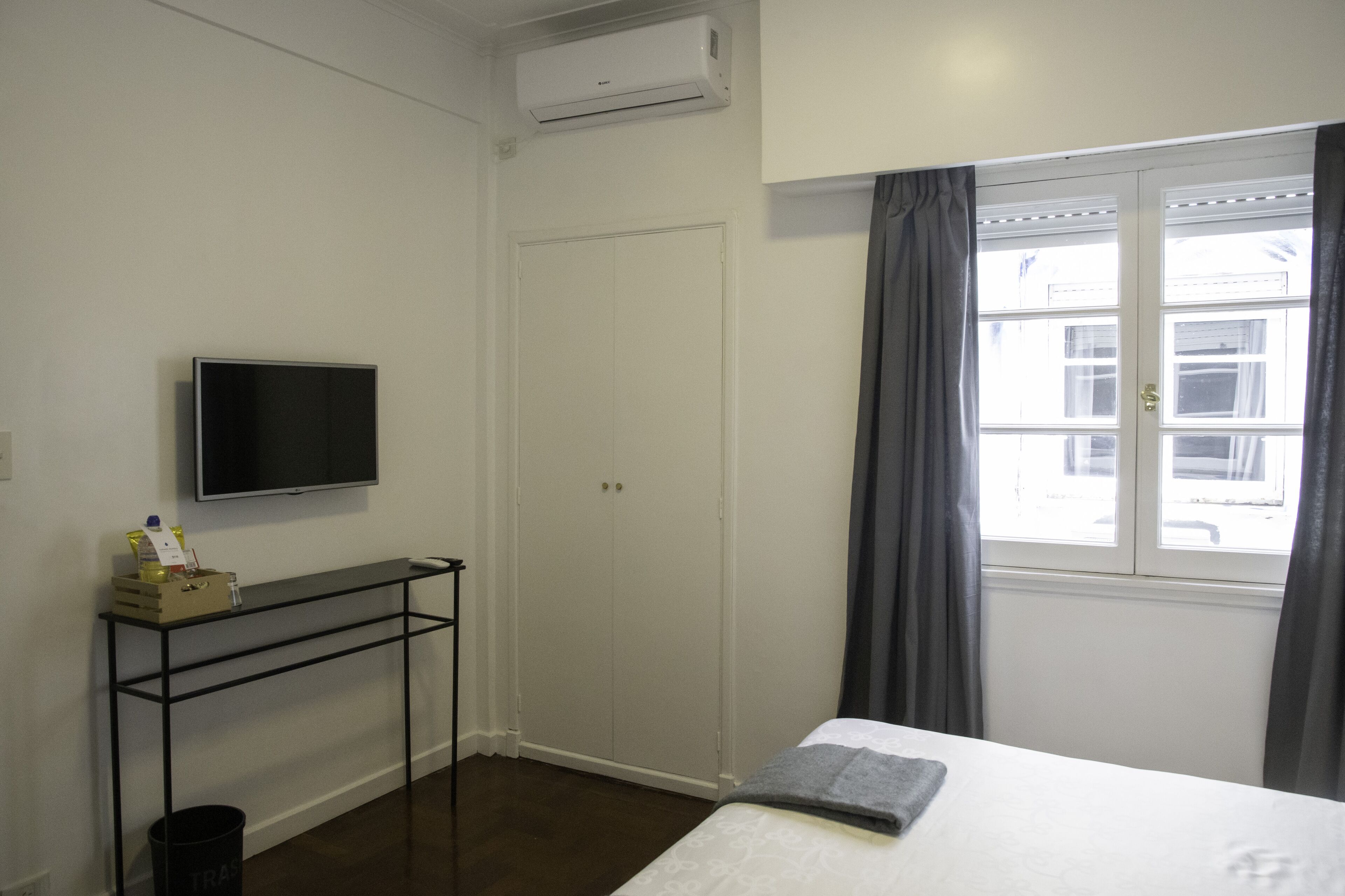 A bedroom with a bed, a TV mounted on the wall, a window with curtains, a wardrobe, and a small table with a box and bottles.