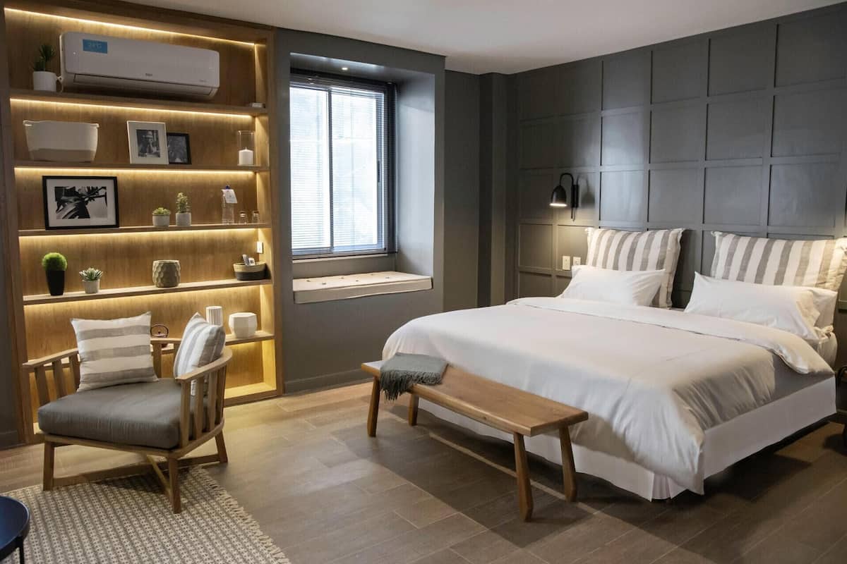 Een moderne slaapkamer met een groot bed, een houten bankje, een stoel, een nachtkastje, een plank met planten en foto’s, en een raam met zonwering.