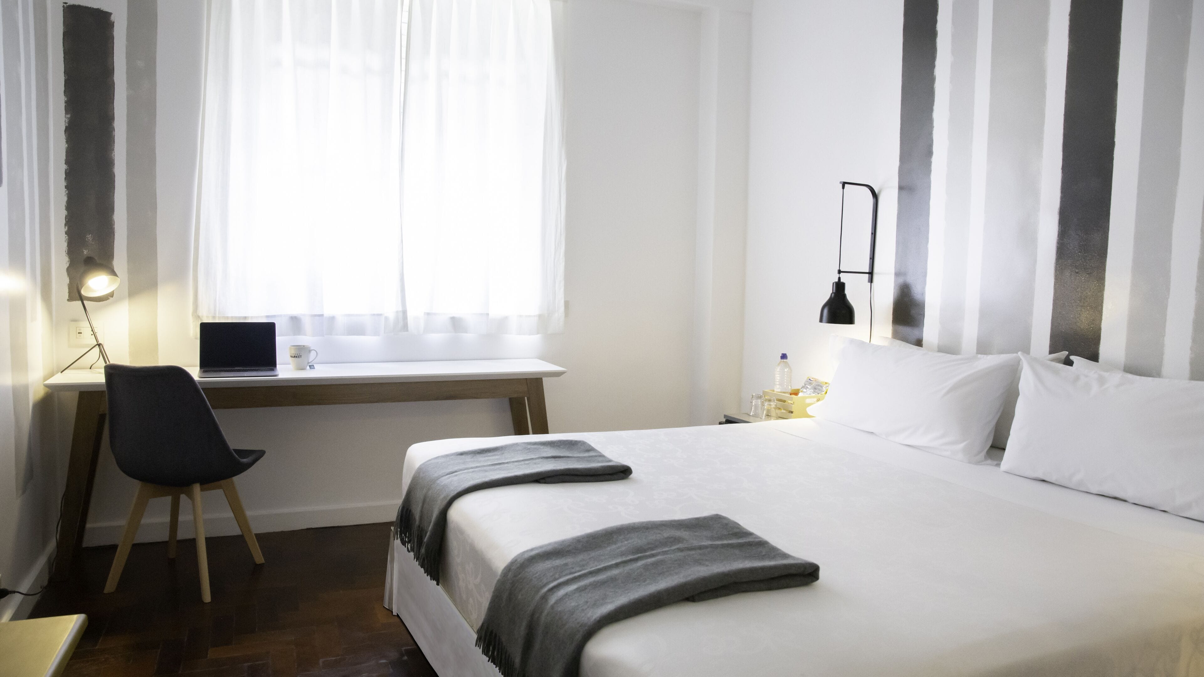 A hotel room with a bed, a desk with a laptop, a chair, a bedside table with a lamp, and a window with sheer curtains.