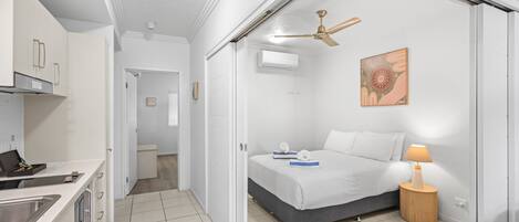 A compact hotel room with a kitchenette, a bed with a grey mattress, a bedside table with a lamp, and a ceiling fan.