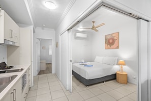 Habitación de hotel compacta con kitchenette, cama con colchón gris, mesita de noche con lámpara y ventilador de techo.
