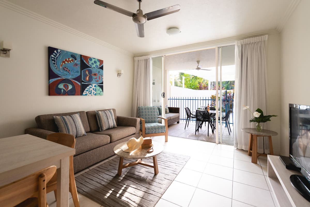 A living room with a sofa, coffee table, and chairs, a dining area with a table and chairs, a TV, and a balcony with a table and chairs.