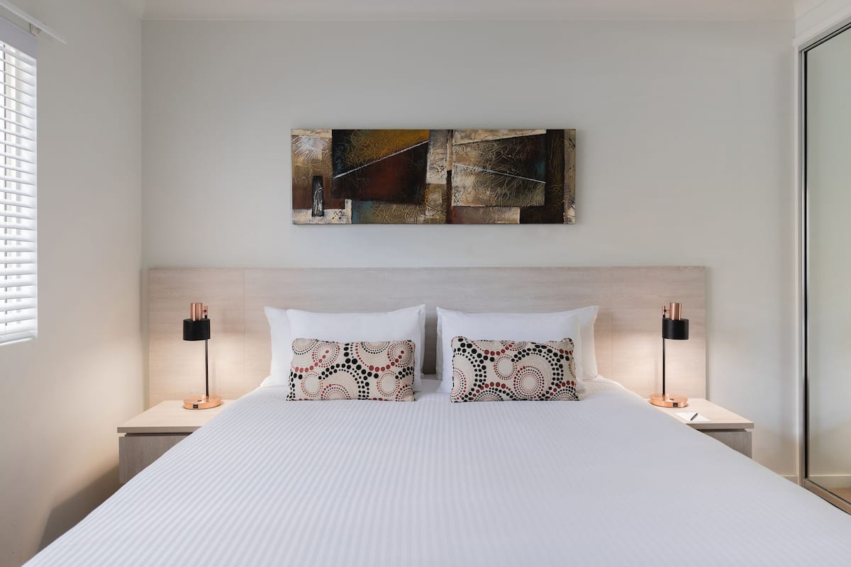 A modern hotel room with a large bed, two bedside tables, a wall-mounted abstract painting, and a window with blinds.