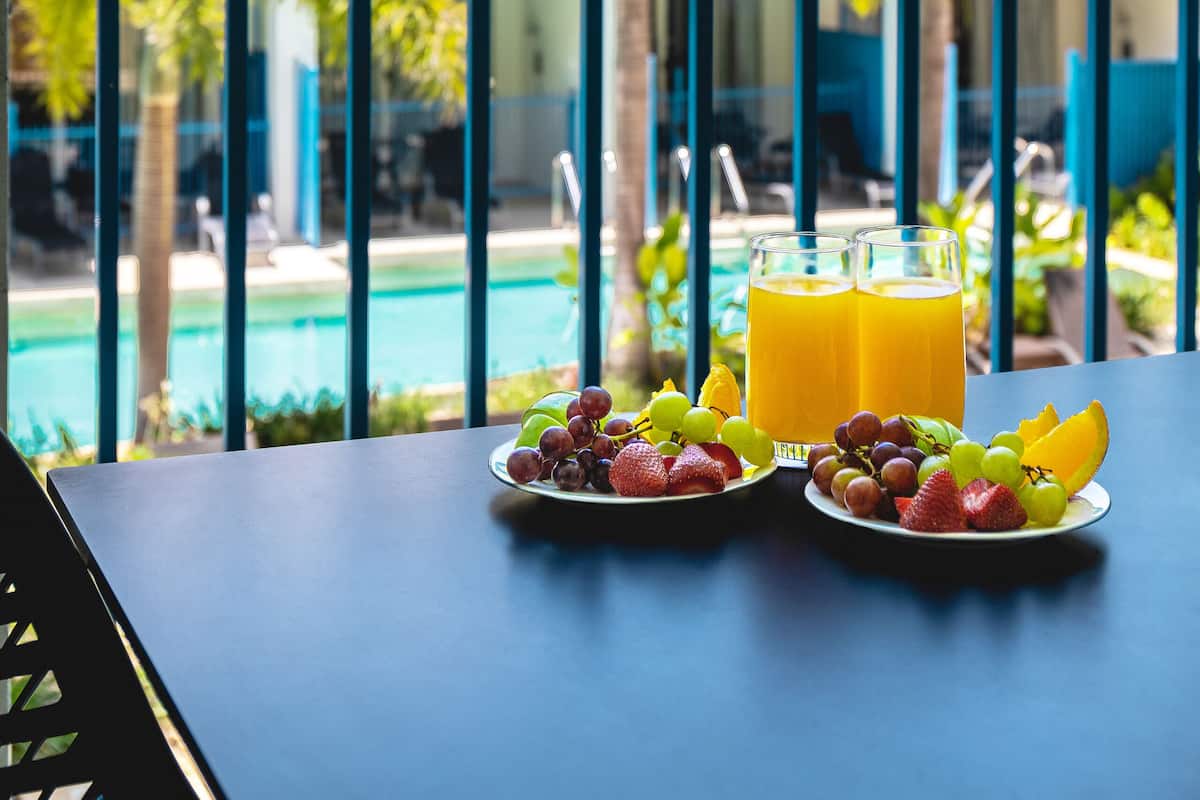 A dining table with plates of fruit and a glass of orange juice, overlooking a pool and palm trees.