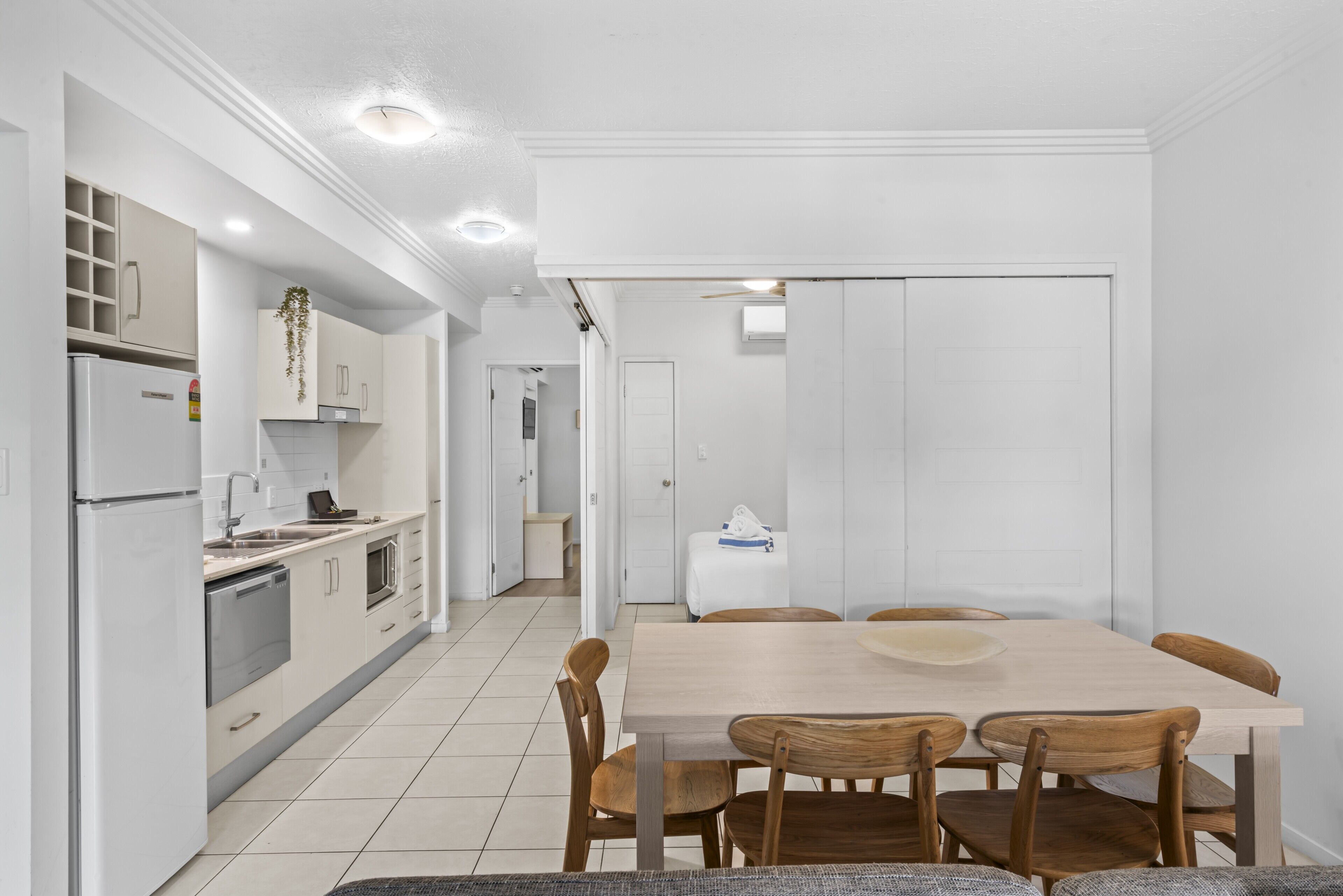 A modern kitchen with white cabinets, a built-in oven, and a refrigerator. There is a dining area with a wooden table and chairs.