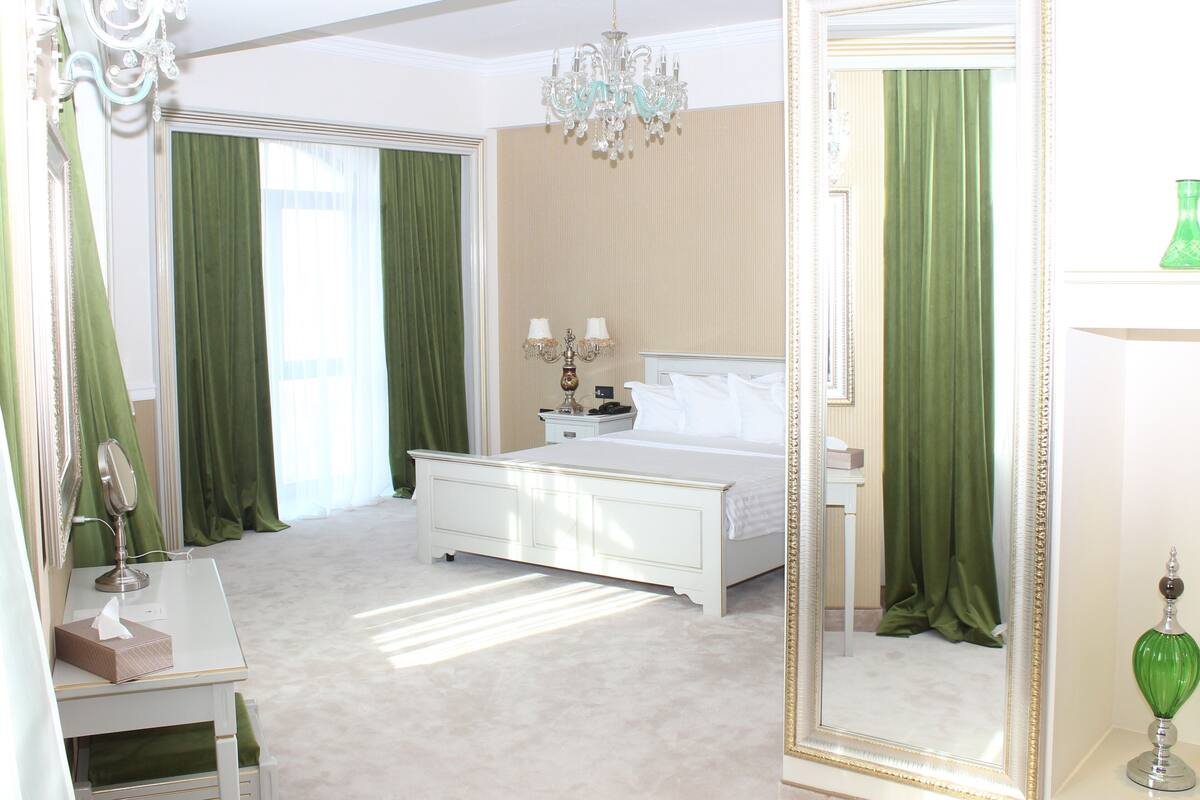 Quarto com cama, mesa de cabeceira, espelho, lustre e cortinas verdes.