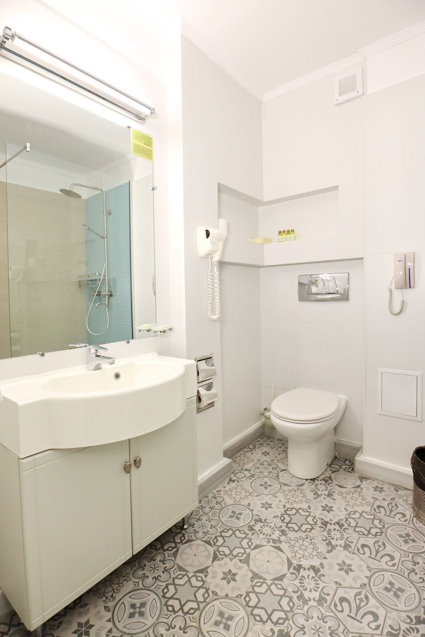 Un baño con lavamanos, espejo y retrete blancos. El piso tiene un diseño y hay un estante pequeño con artículos de aseo.