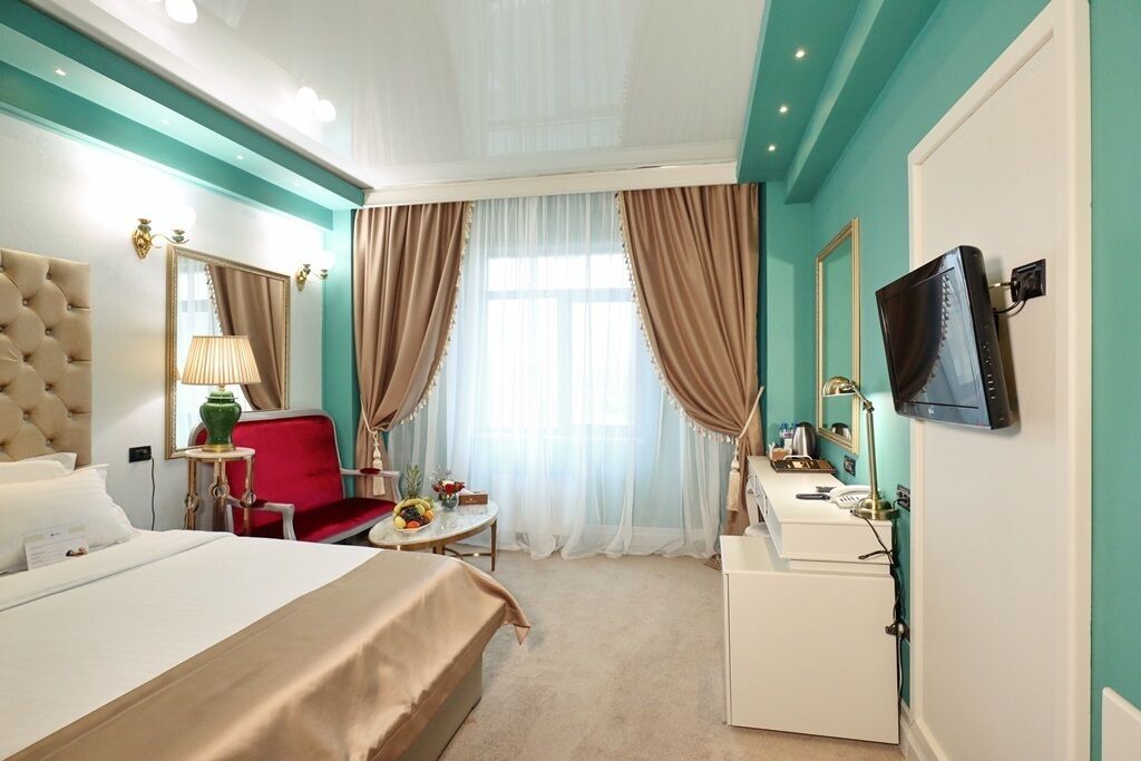 A hotel room with a large bed, a red chair, a small table with fruit, a desk with a lamp, and a TV on the wall.