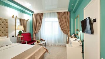 A hotel room with a large bed, a red chair, a small table with fruit, a desk with a lamp, and a TV on the wall.