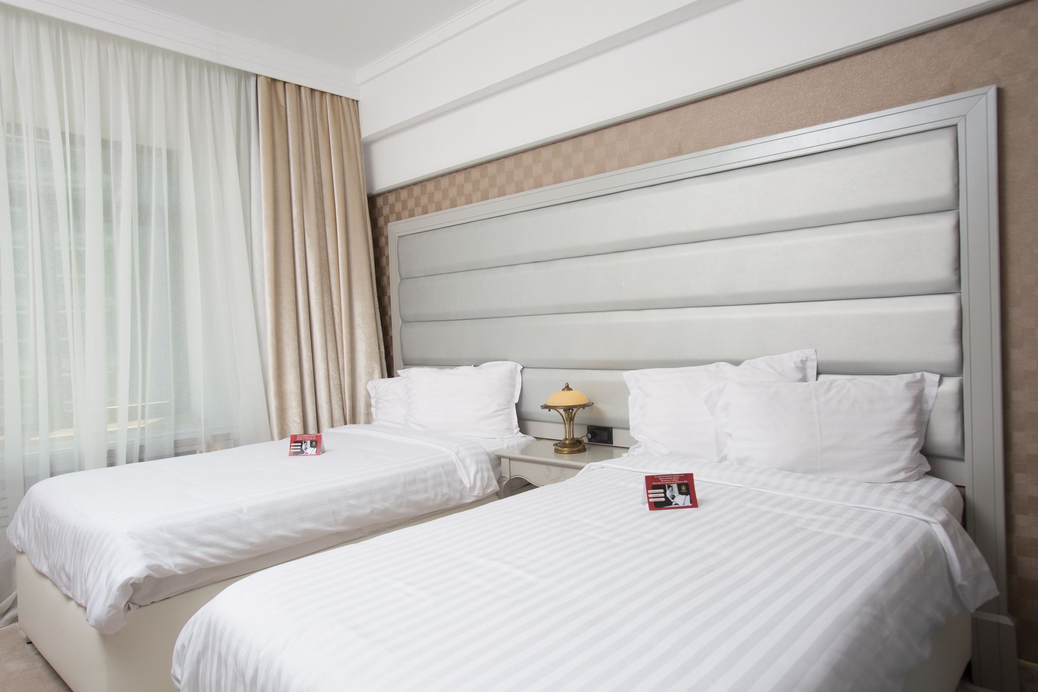 Habitación de hotel con dos camas individuales, una mesita de noche con lámpara y un ventanal con cortinas traslúcidas.