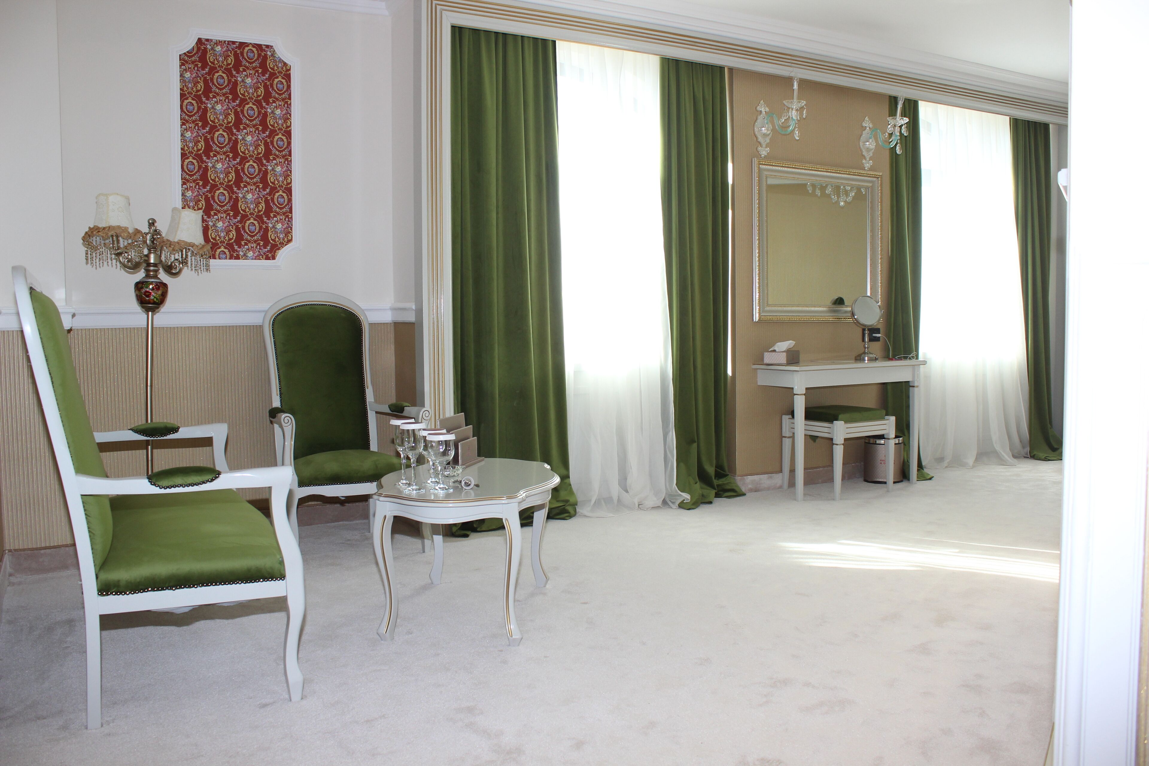 A room with a green upholstered chair, a small table with a mirror, and a white vanity with a green curtain.