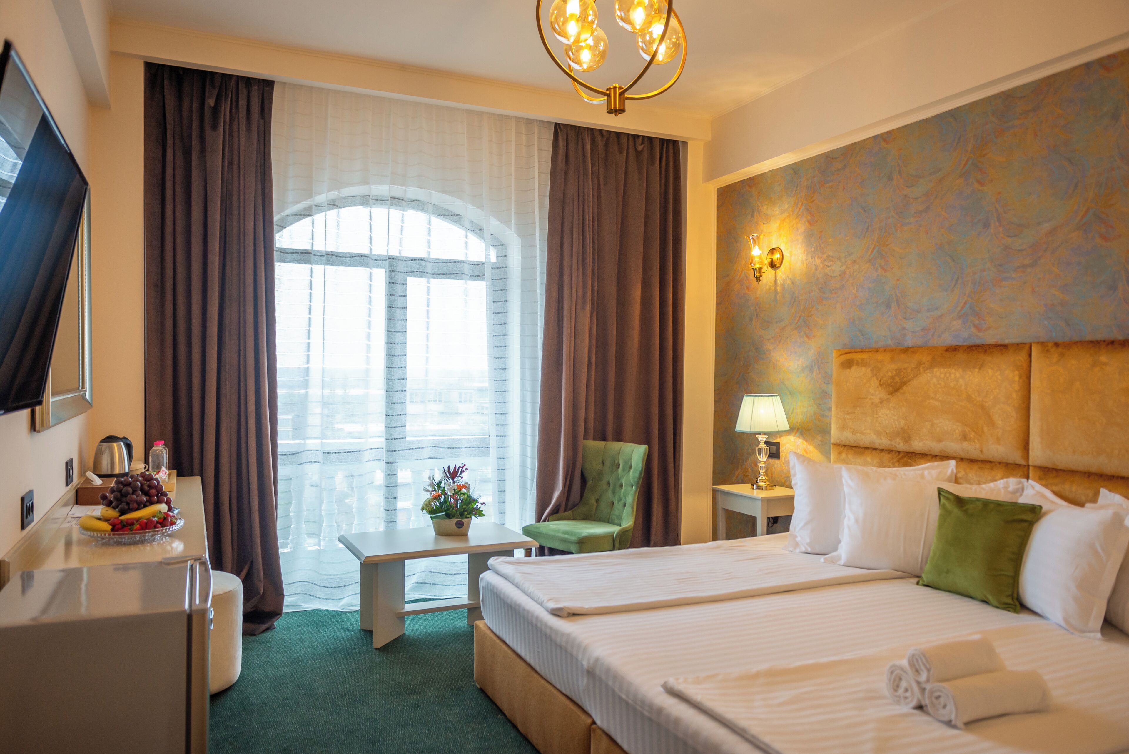 Habitación de hotel con una cama, un escritorio con frutero, una silla, un televisor y una ventana con cortinas.