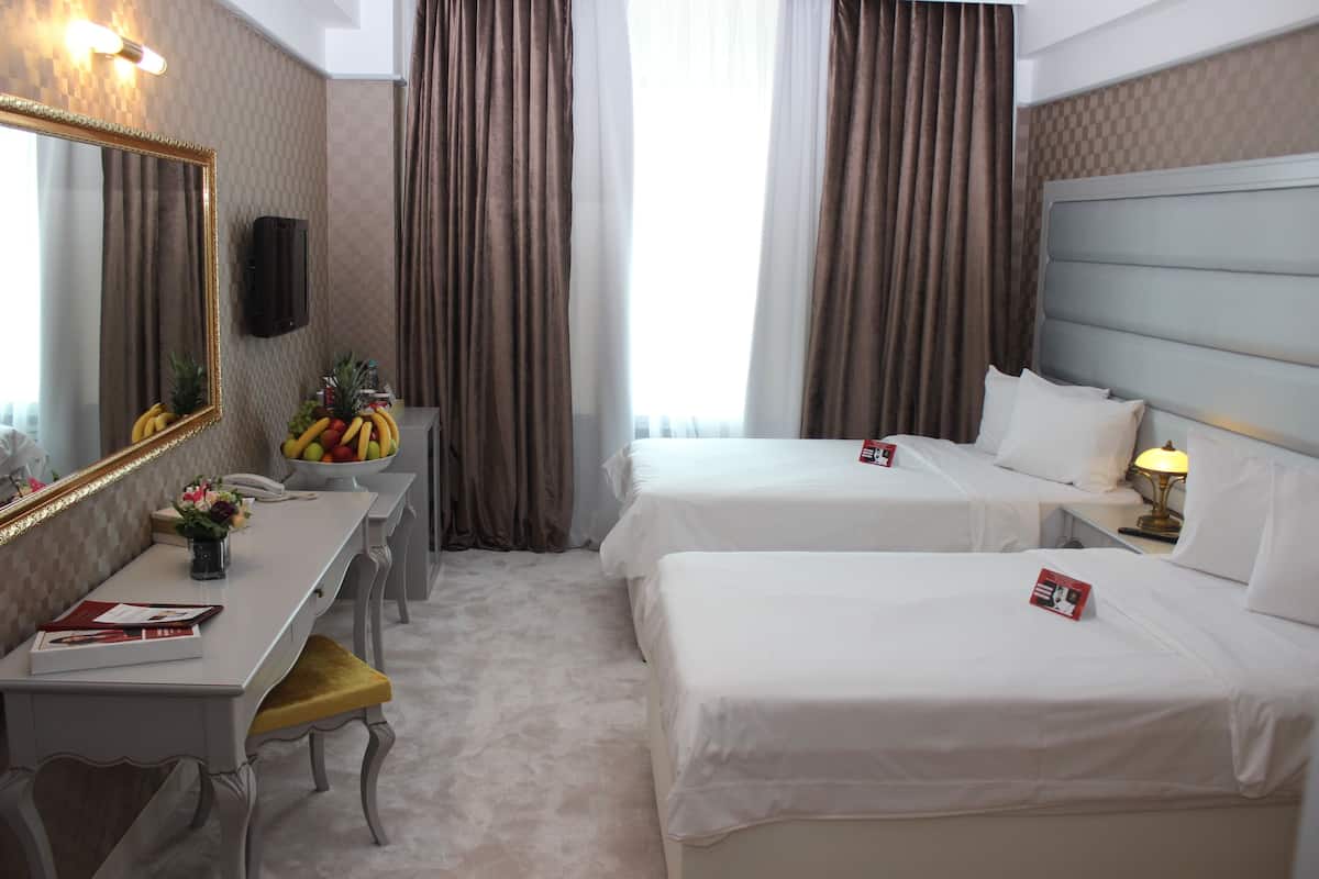 Quarto de hotel com duas camas, uma escrivaninha com espelho, uma cesta de frutas e uma pequena mesa com abajur.