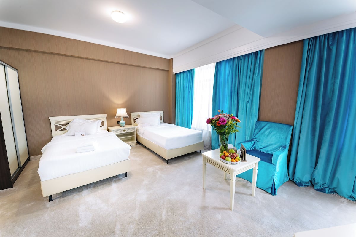 Quarto de hotel com duas camas, uma mesa de cabeceira com um vaso de flores e vista para o mar através de grandes janelas com cortinas azuis.