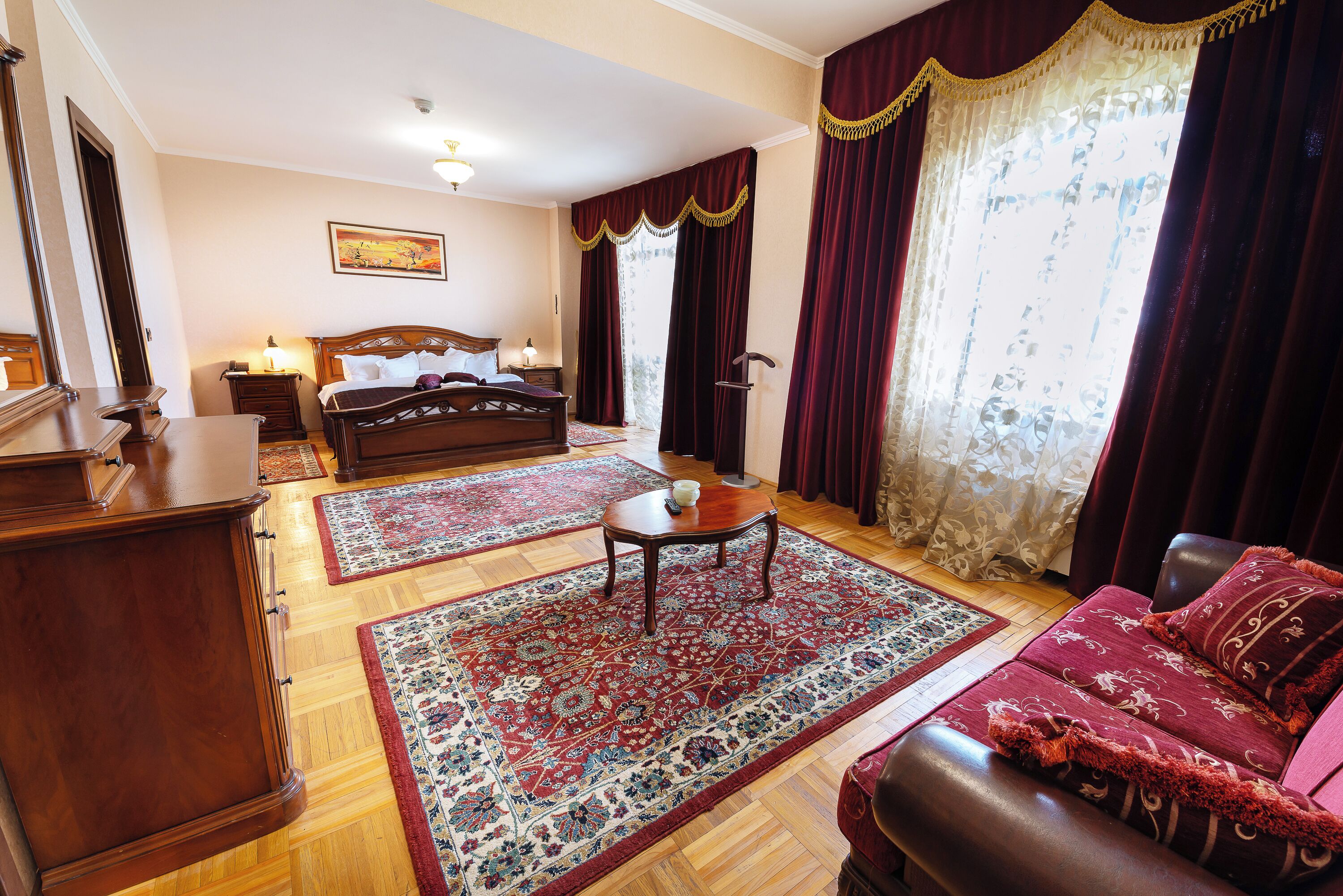 Quarto de hotel com cama, sofá, mesa de centro, mesa de cabeceira, abajur e uma grande janela com cortinas.