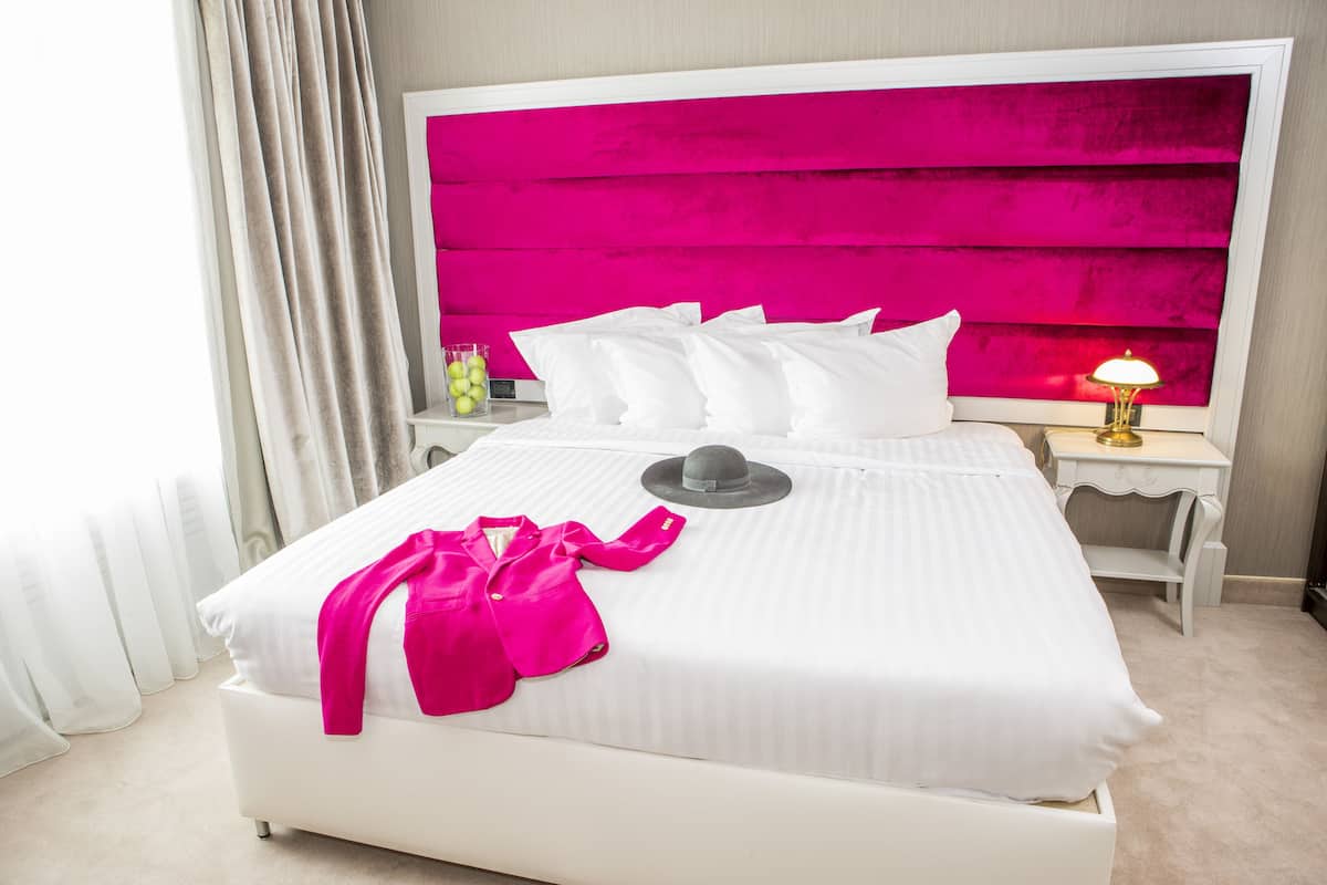 Quarto de hotel com uma cama grande, roupa de cama branca, cabeceira rosa vibrante, mesa de cabeceira com abajur e criado-mudo com um vaso de flores.