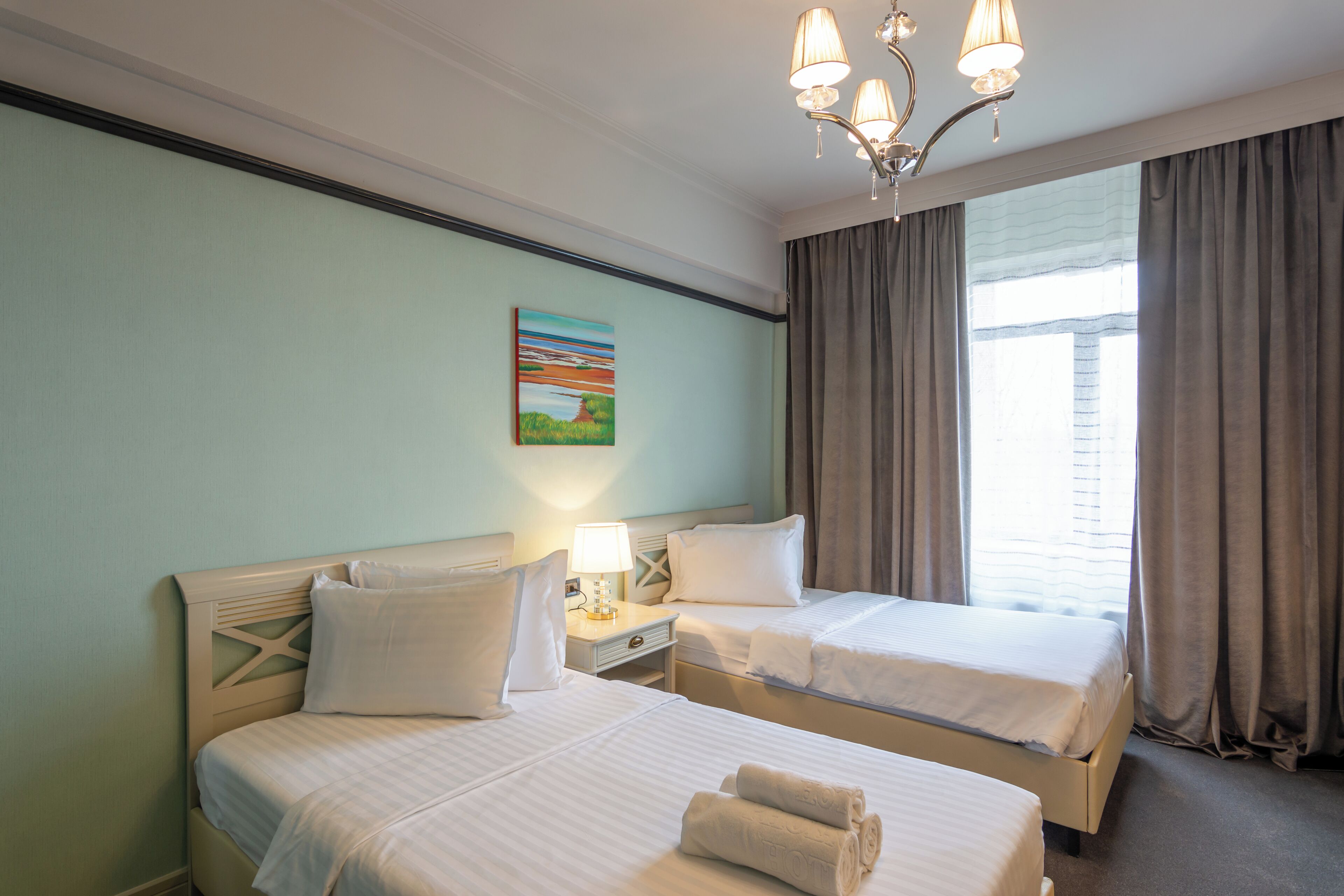 A hotel room with two beds, a nightstand with a lamp, a painting on the wall, and a window with curtains.