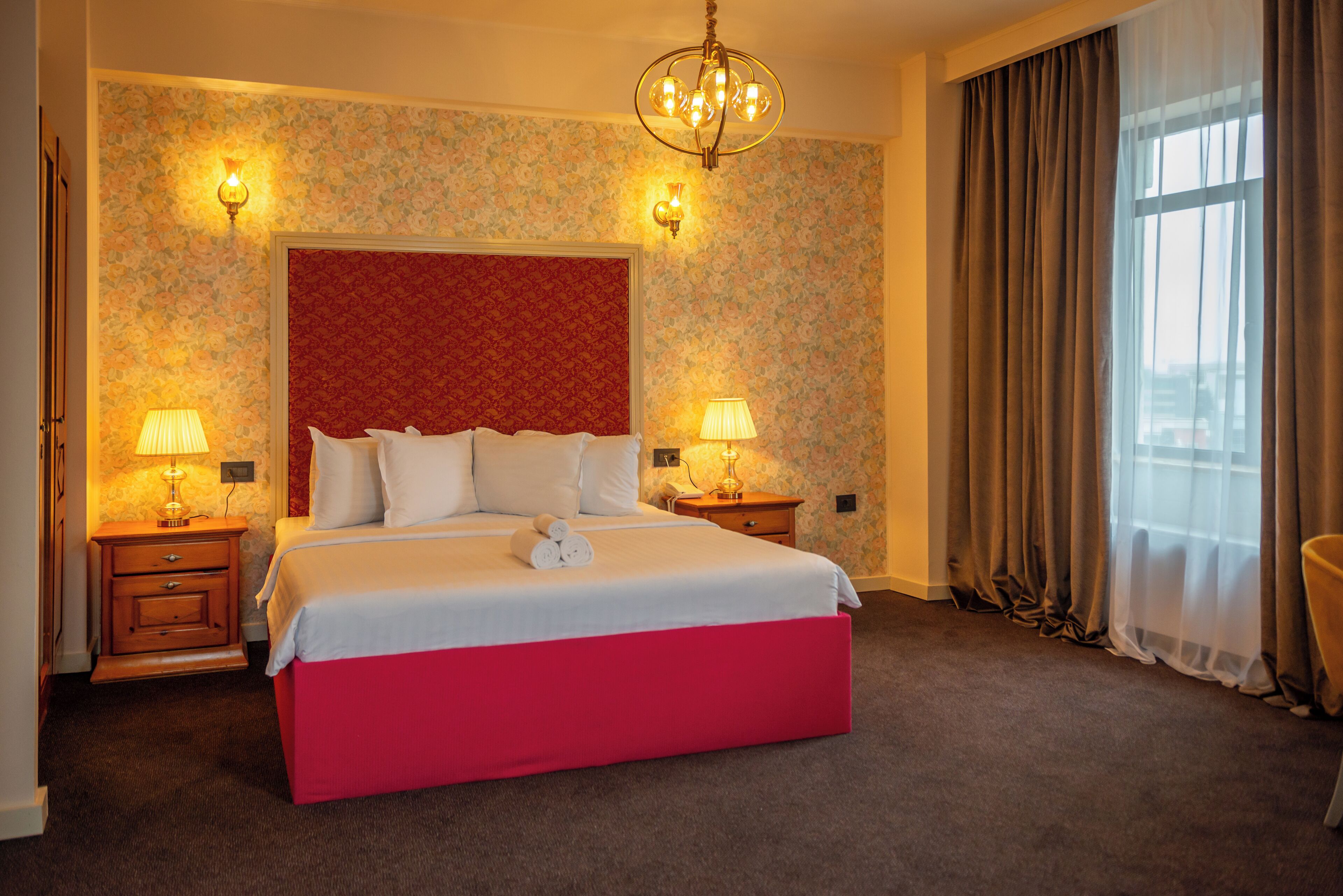 Une chambre d’hôtel avec un grand lit, deux tables de chevet avec des lampes, un lustre et une vue sur la ville par la fenêtre.