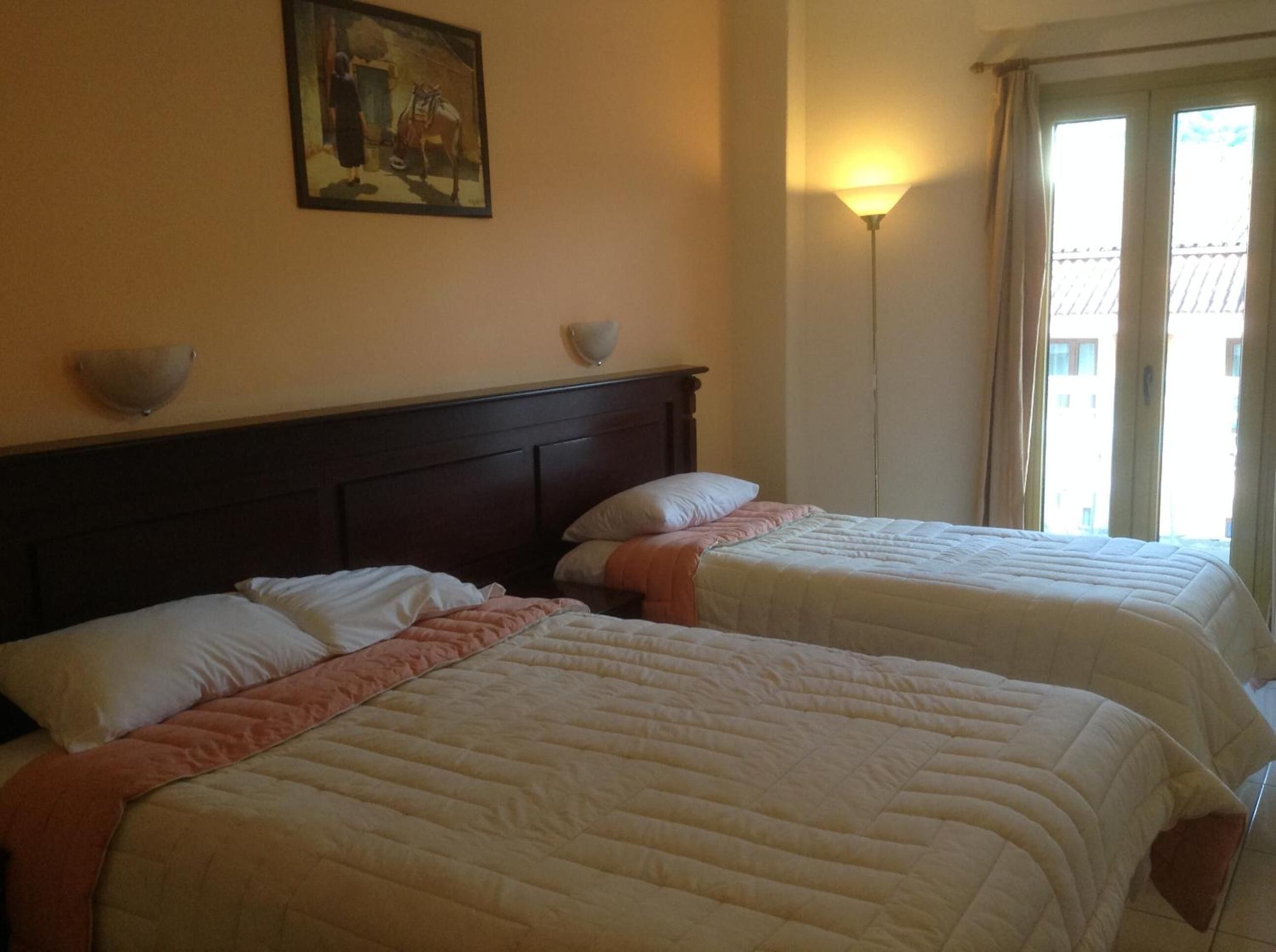 Dos camas individuales con cabeceros, una lámpara de pared, un cuadro y una ventana con cortinas.