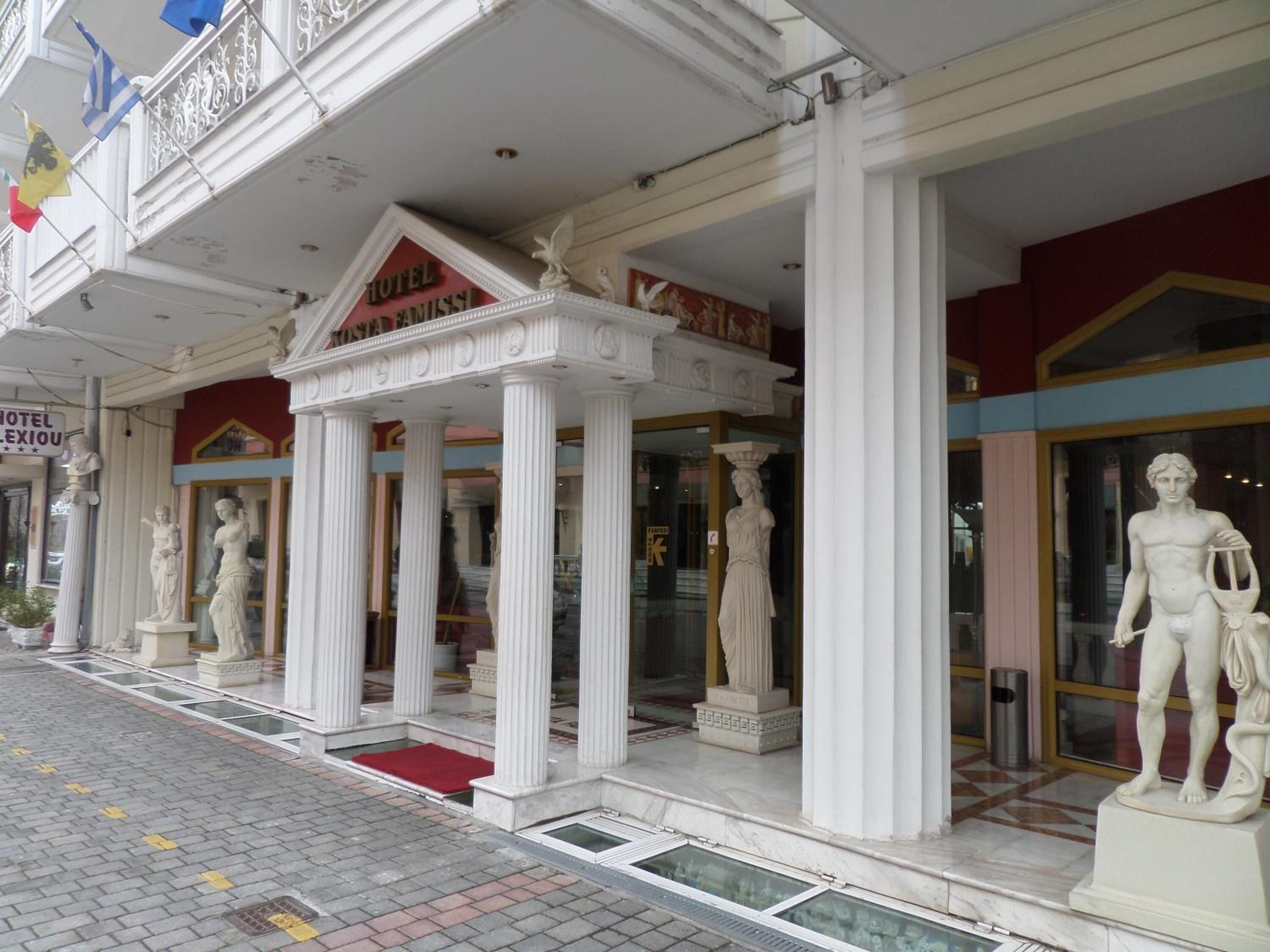Entrada de un hotel con columnas clásicas, un toldo rojo y una estatua de un hombre sosteniendo una lira.