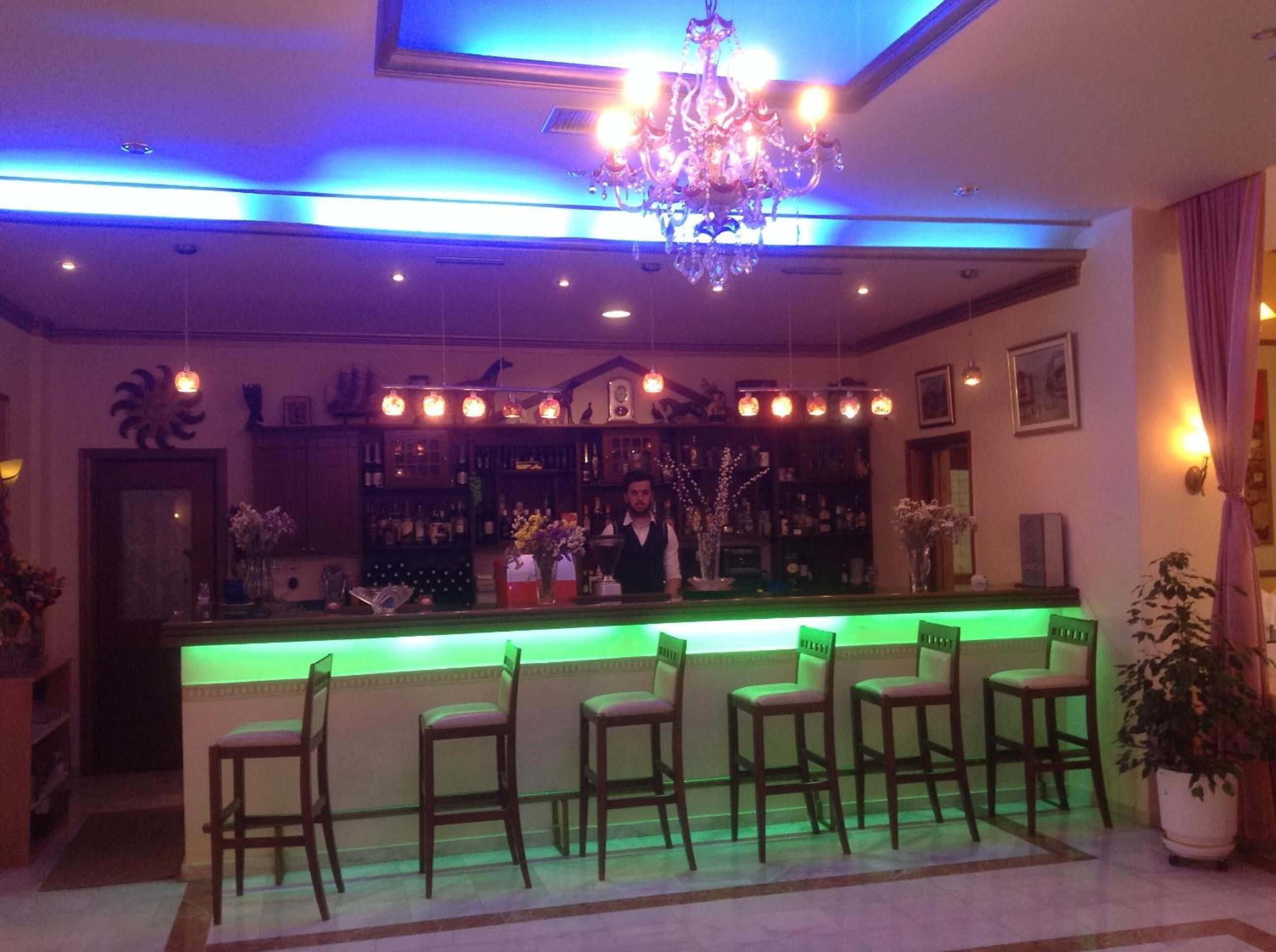 A bar area with a green-lit bar counter, multiple bar stools, a chandelier, and a bartender.