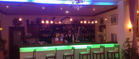 A bar area with a green-lit bar counter, multiple bar stools, a chandelier, and a bartender.