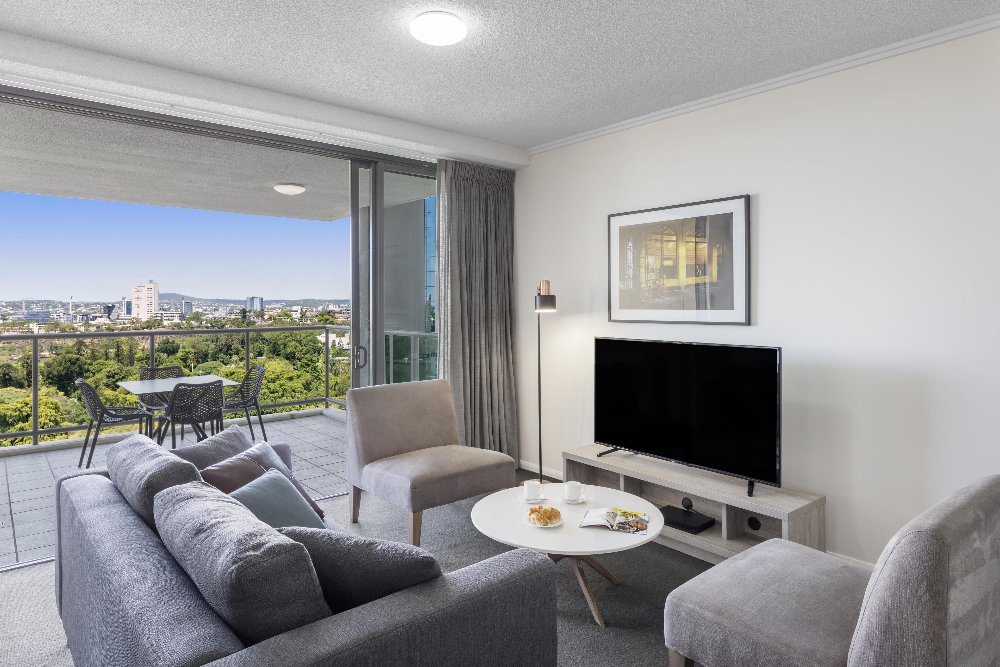 A modern living room with a sofa, armchair, and TV on a stand, with a balcony and city view.